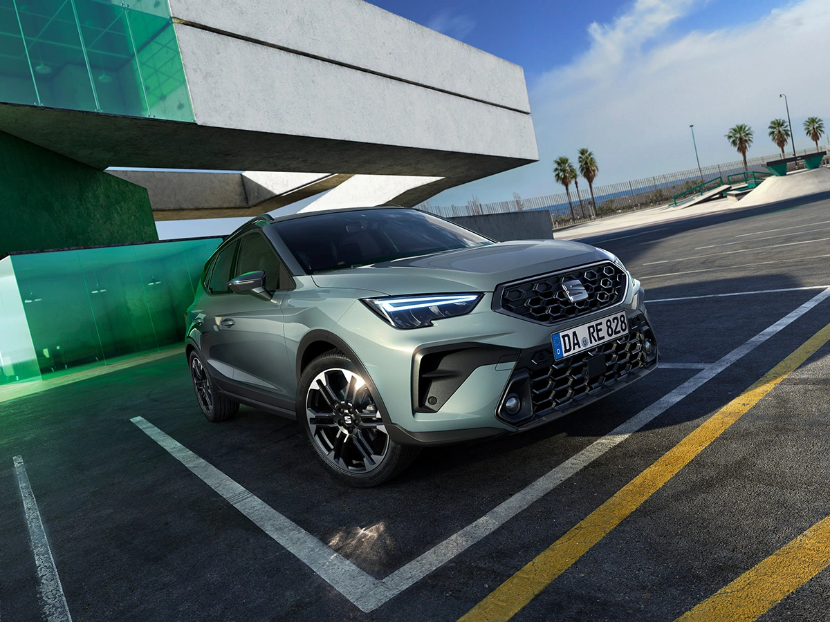 SEAT Arona in der Farbe Oniric Lake vor grünem Glasgebäude auf leerem Parkplatz